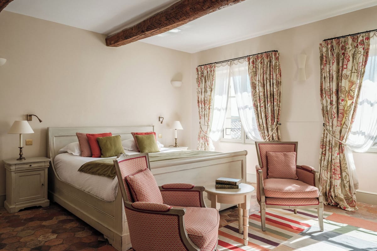 Un dormitorio con una cama, dos sillas, una mesita y una ventana con cortinas.