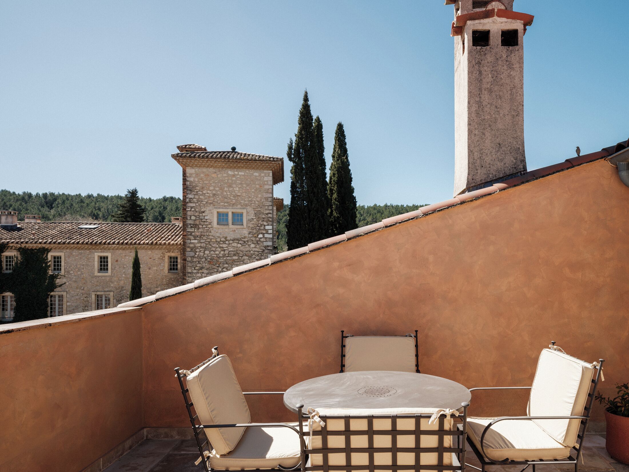 A terrace with a round table and two chairs, a stone building in the background, and a clear sky.