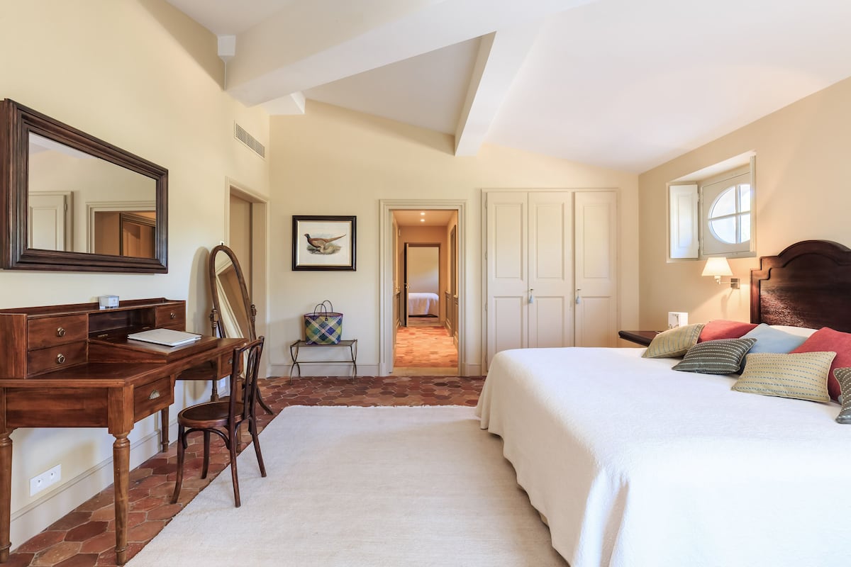 Un dormitorio con una cama grande, un escritorio, una silla y un espejo. Hay una puerta que da a otra habitación.
