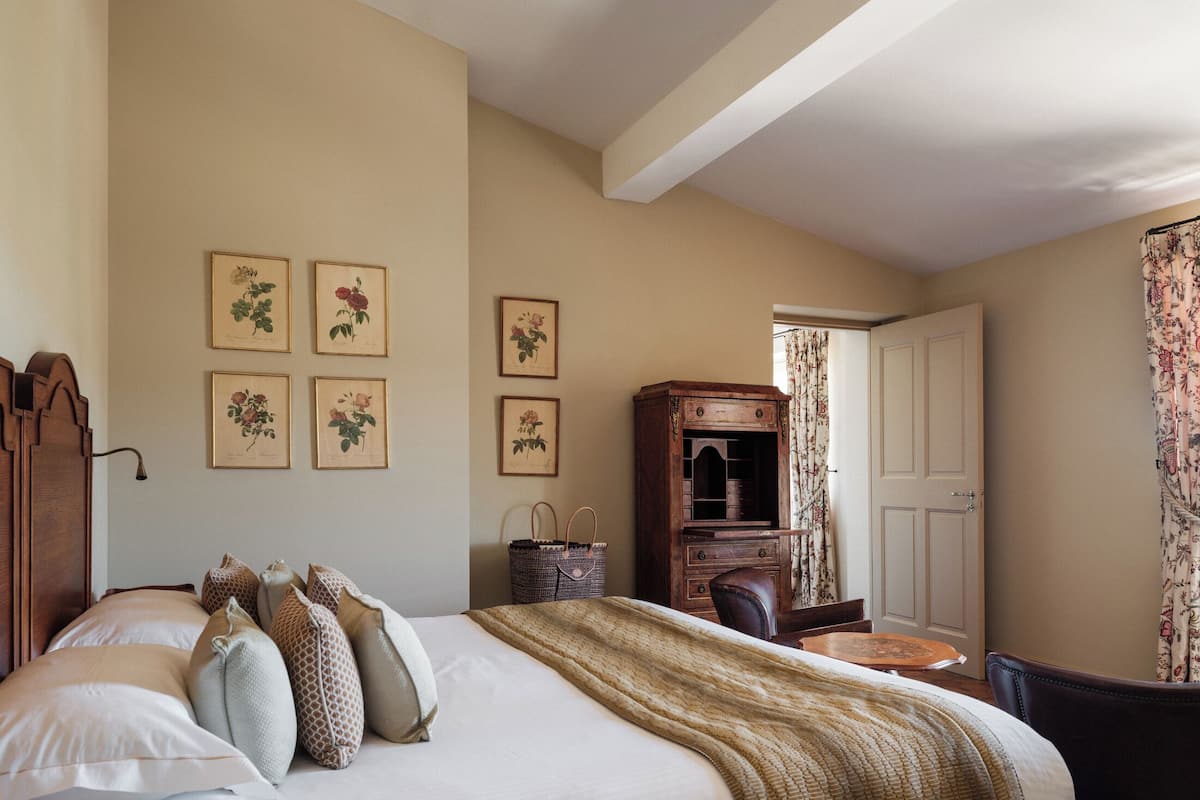 Un dormitorio con cama, almohadas, un aparador de madera, una silla e impresiones botánicas enmarcadas en la pared.