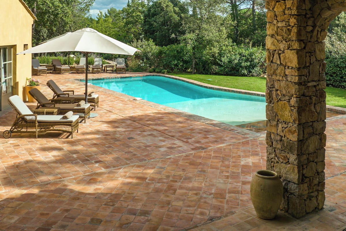 Una piscina con suelo de baldosas, sillones de descanso y un pilar de piedra.