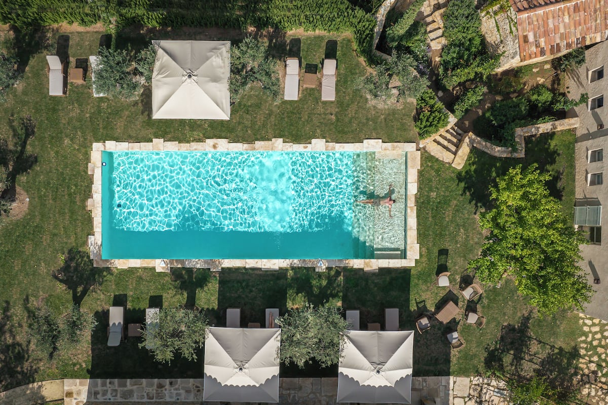 Vista aérea de una piscina rectangular con un borde de baldosas, dos sombrillas y un muro de piedra.