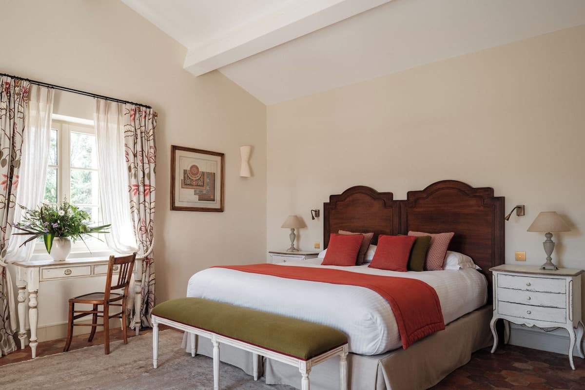 Un dormitorio con cama, mesitas de noche, escritorio, silla, banco y ventana con cortinas.