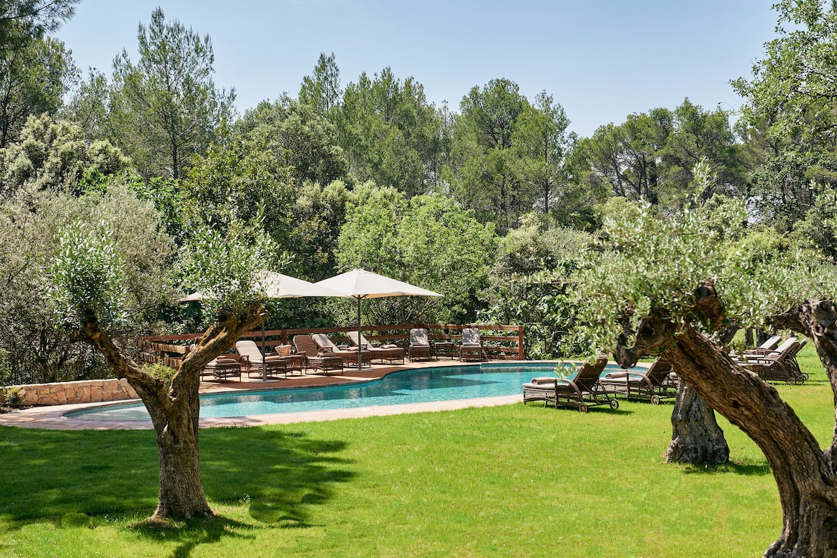 Un área de piscina con sillas de descanso y sombrillas, rodeada de árboles.