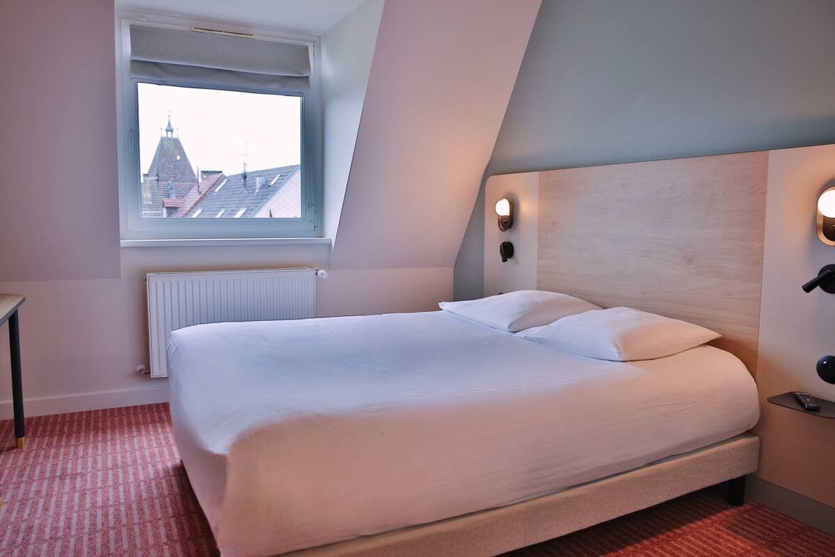 Hotelkamer met een groot bed, een raam met uitzicht op gebouwen, een radiator en een nachtkastje met een lamp.