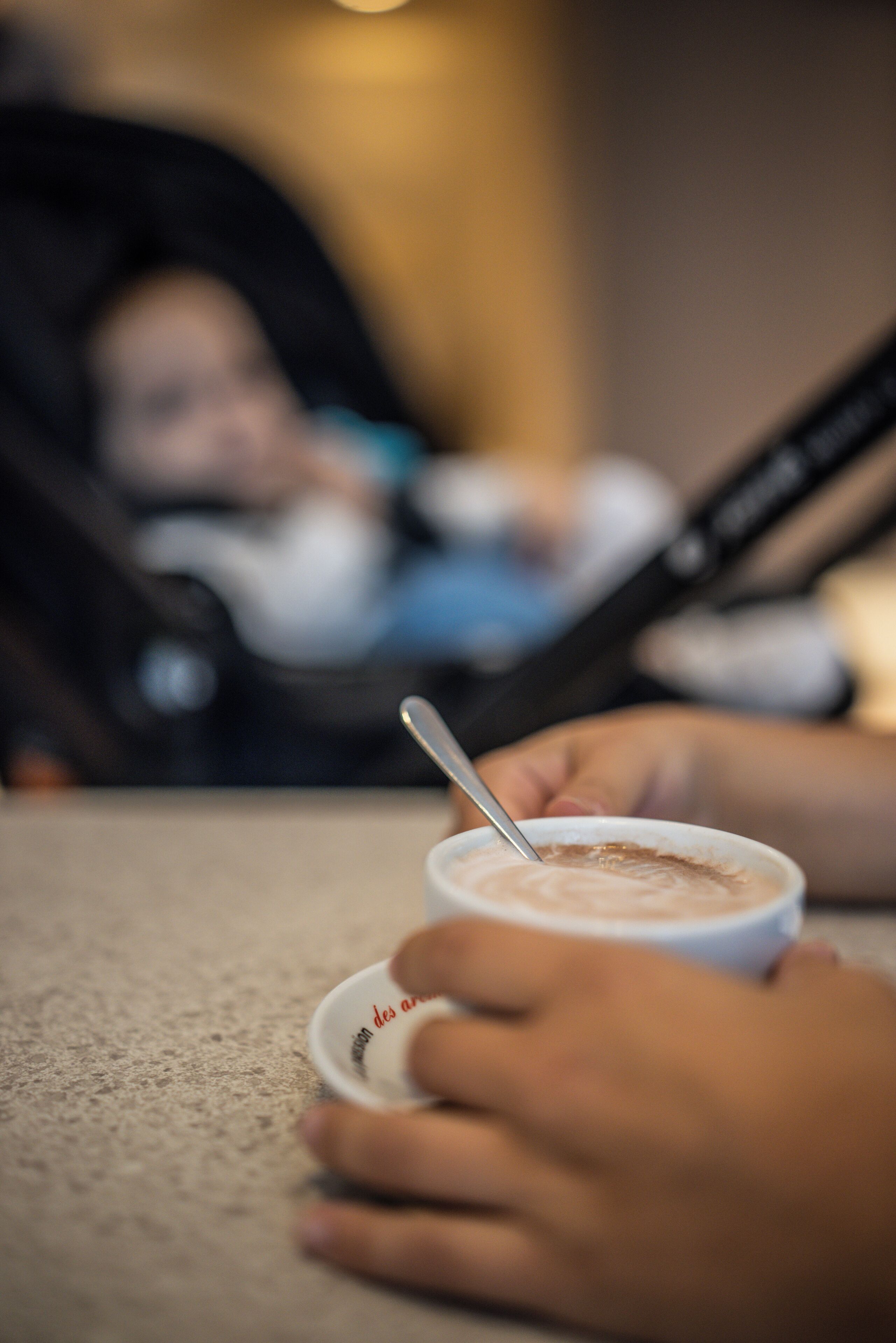 A close-up of a hand holding a cup with a spoon, with a blurred background of a stroller.