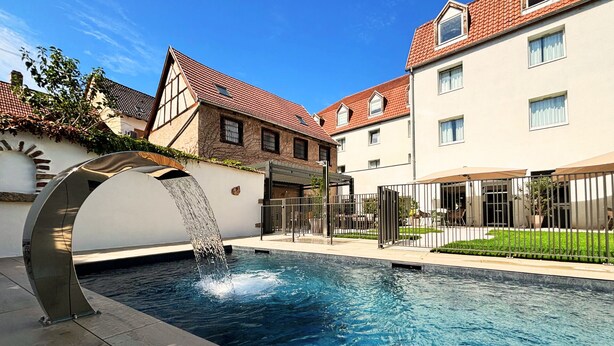 A swimming pool with a water feature, surrounded by buildings with red-tiled roofs and a covered outdoor seating area.