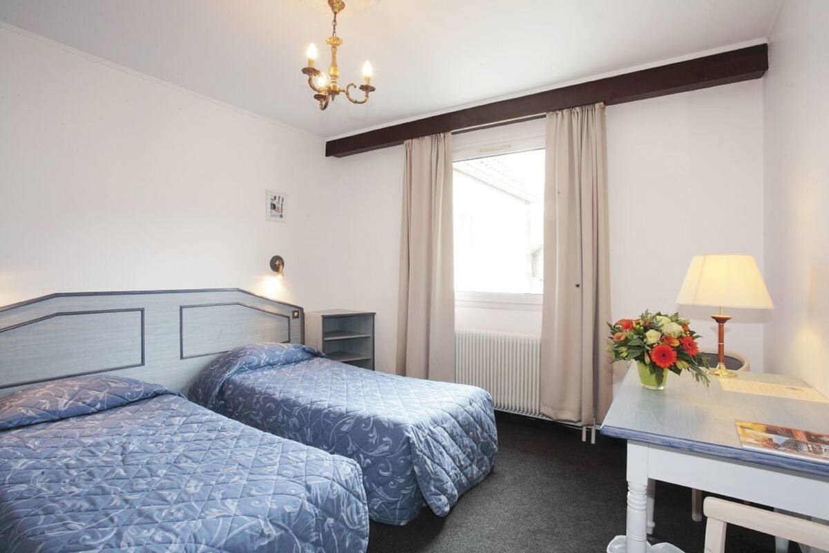Hotelkamer met twee bedden, een bureau, een lamp, een vaas met bloemen, een radiator en een kroonluchter.
