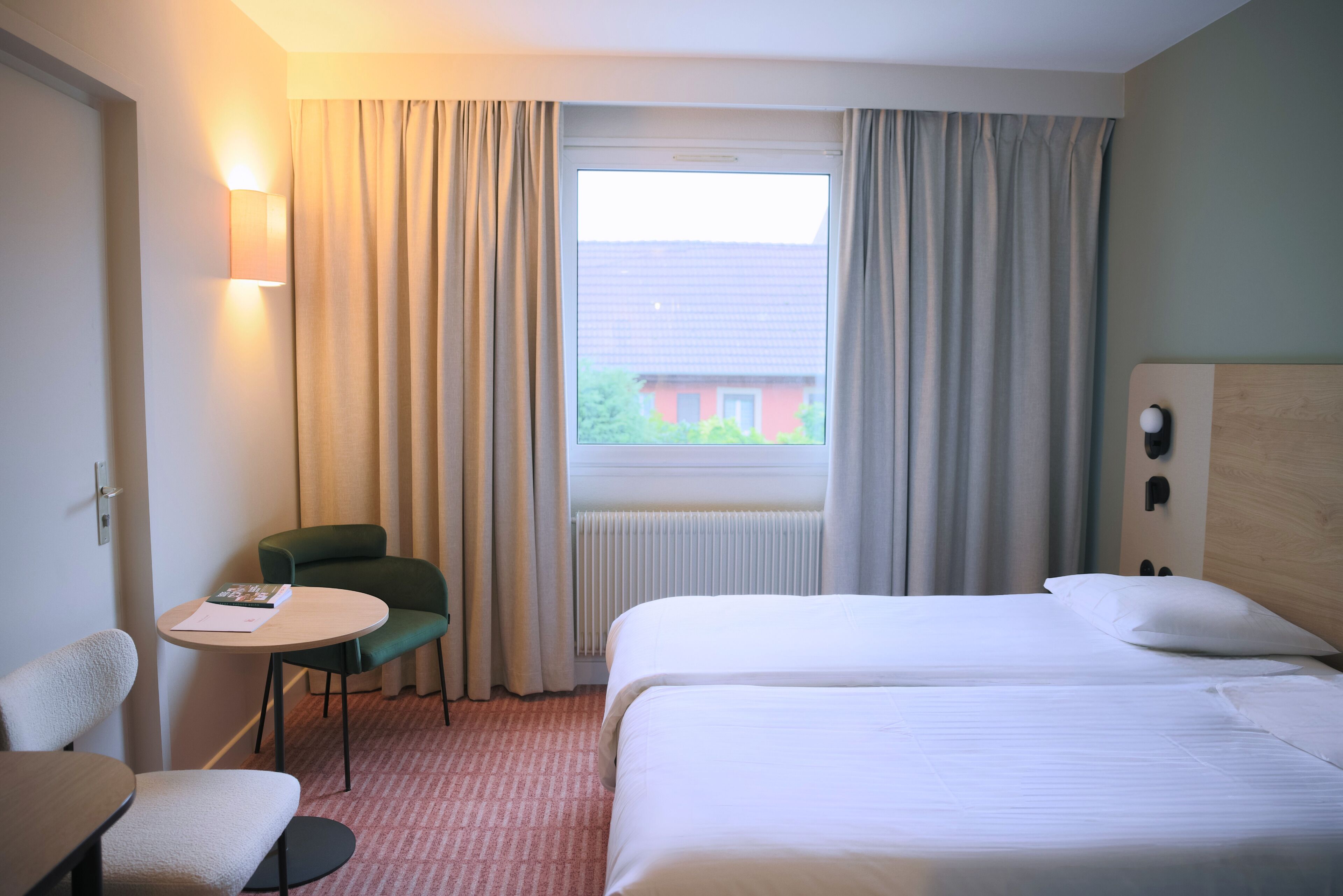 A hotel room with two beds, a small table, a chair, and a window with curtains.