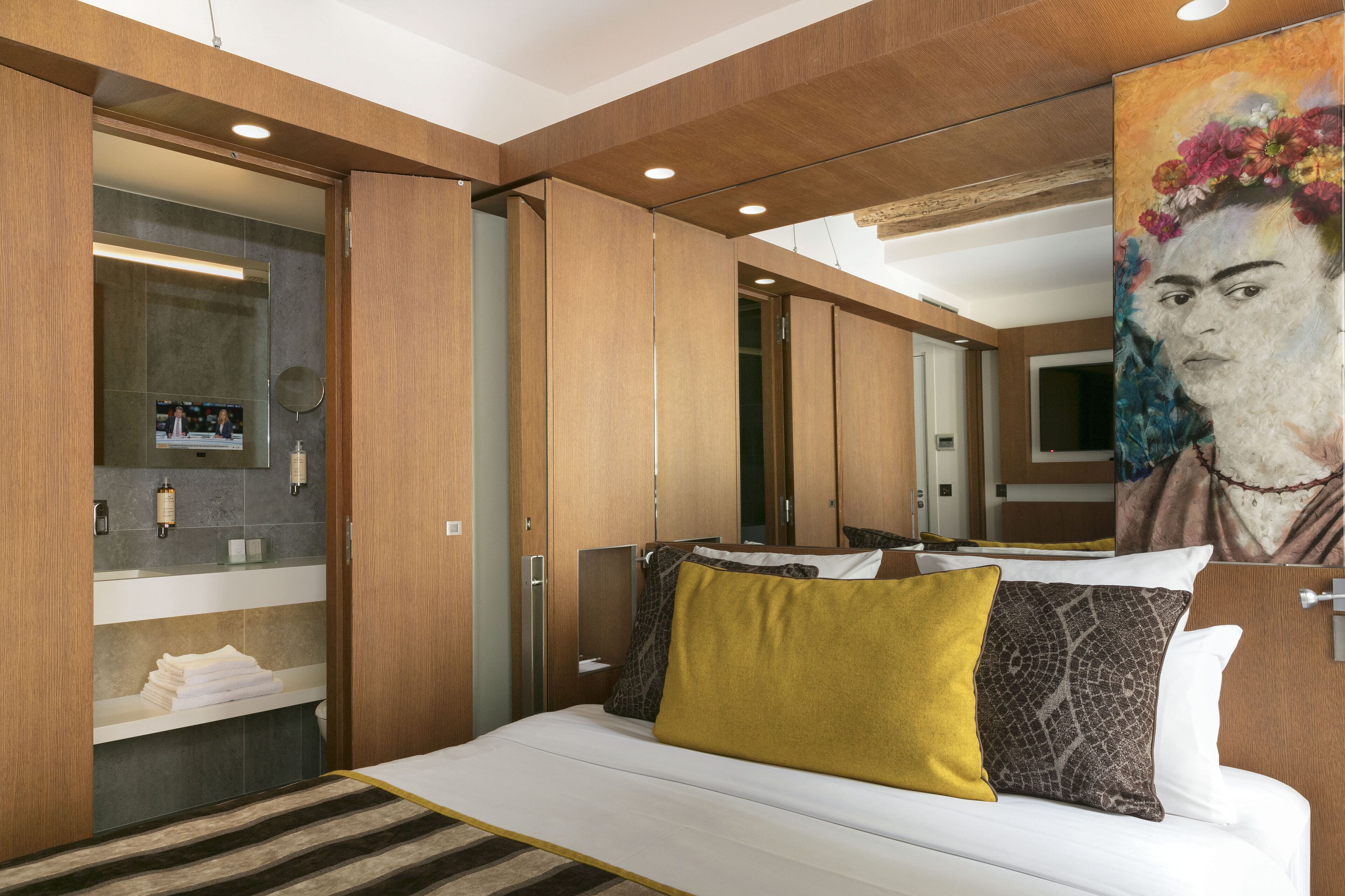 A hotel room with a bed, a mirror, a bathroom with a sink, and a painting on the wall.