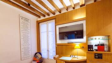 A compact hotel room with a wooden ceiling, a large window, a flat-screen TV, a small desk with a lamp, and a kitchenette area.