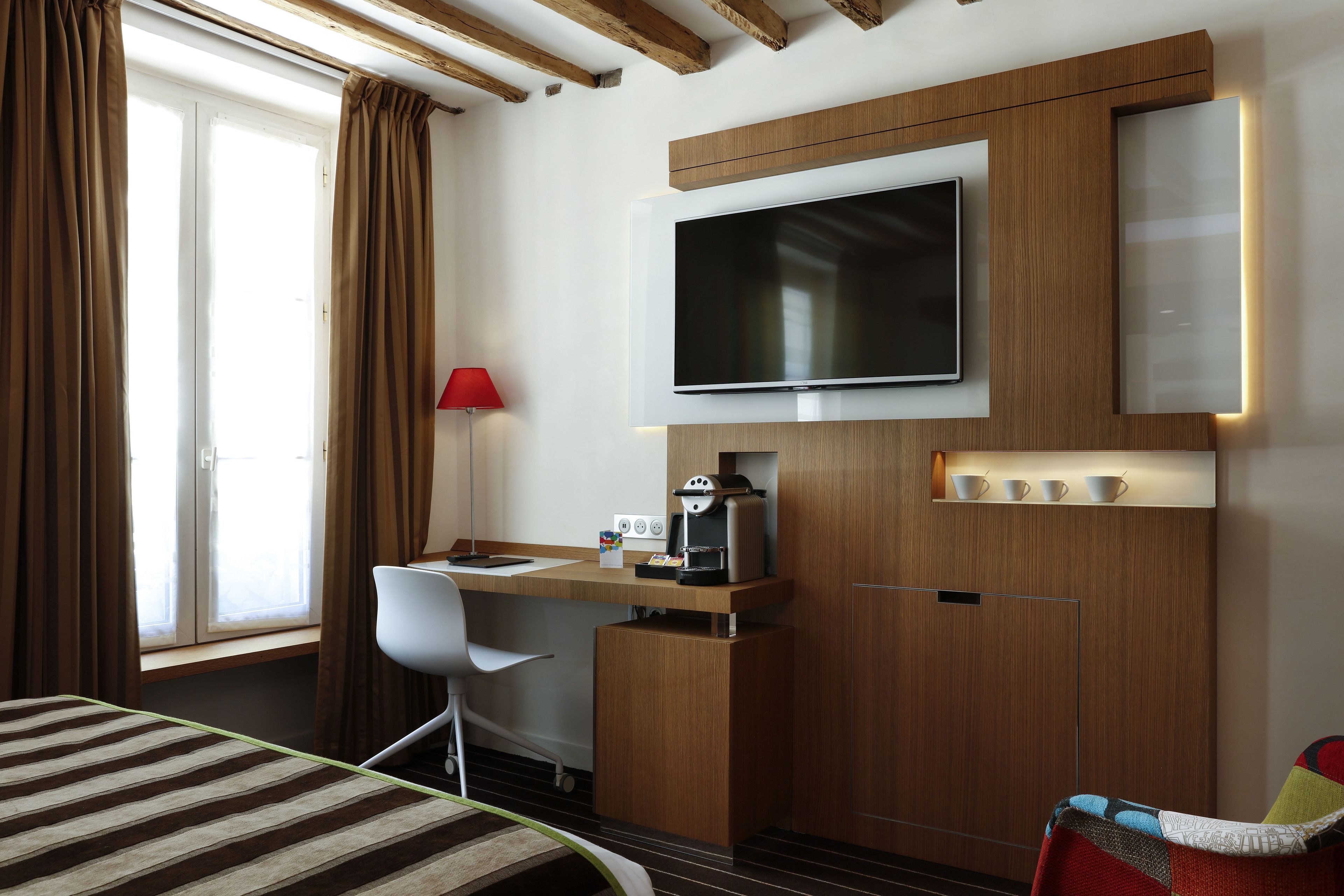 Habitación de hotel con cama, escritorio y silla, televisor en la pared, cafetera y ventana con cortinas.