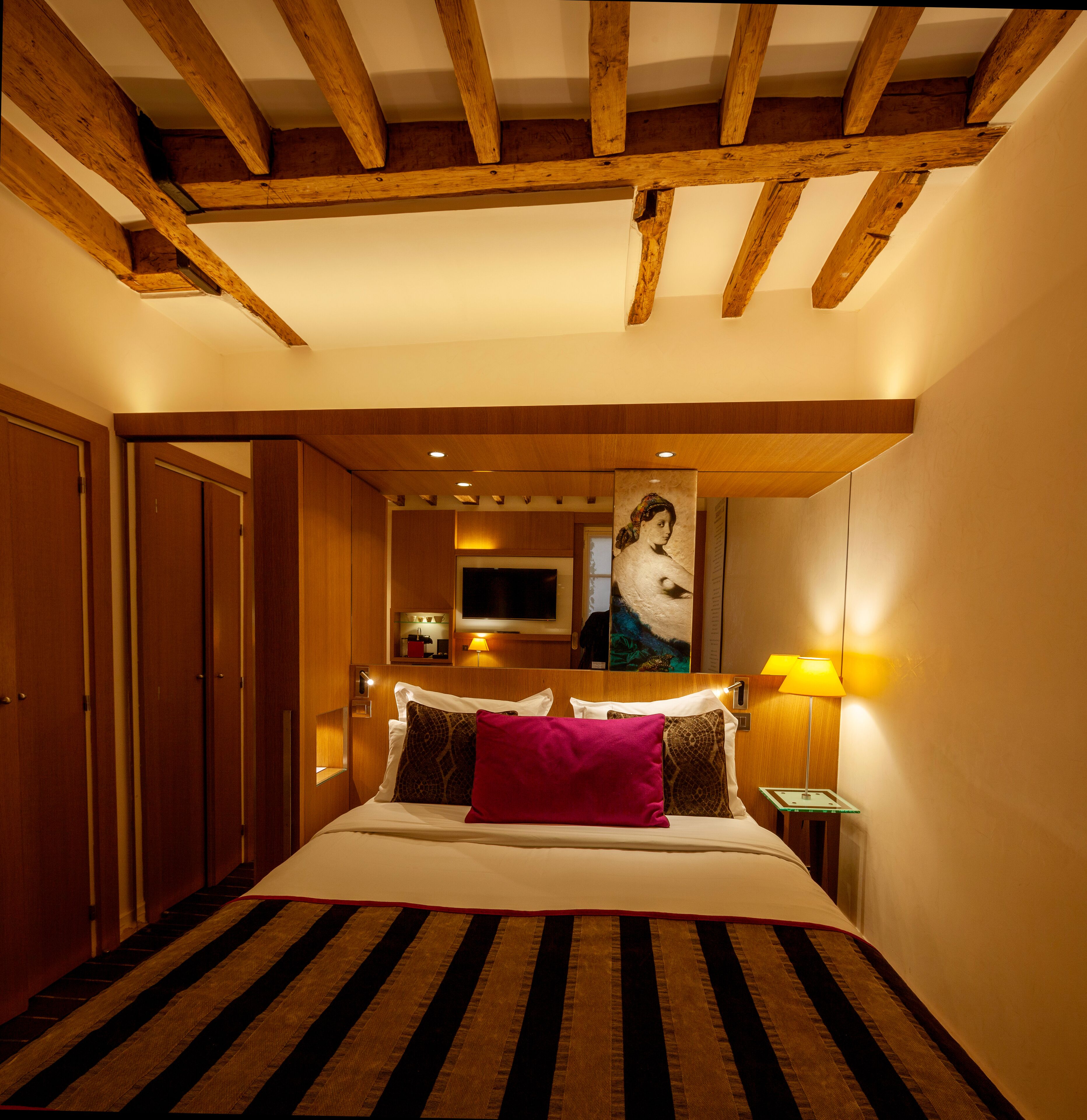 A bedroom with a wooden ceiling, a bed with striped bedding, a painting on the wall, and a bedside lamp.