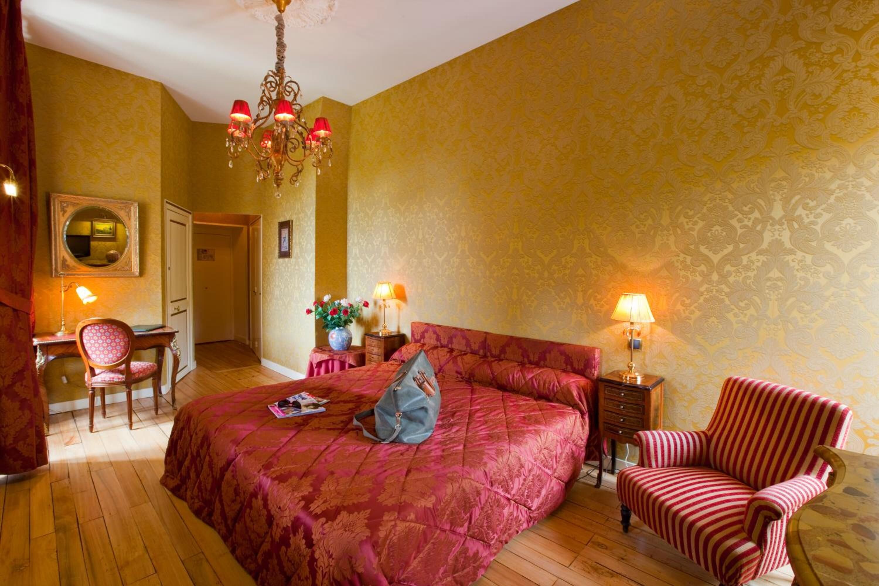 Een ruime slaapkamer met een groot bed, een bureau en een stoel. Er is een kroonluchter, een spiegel en een deur naar een andere kamer.