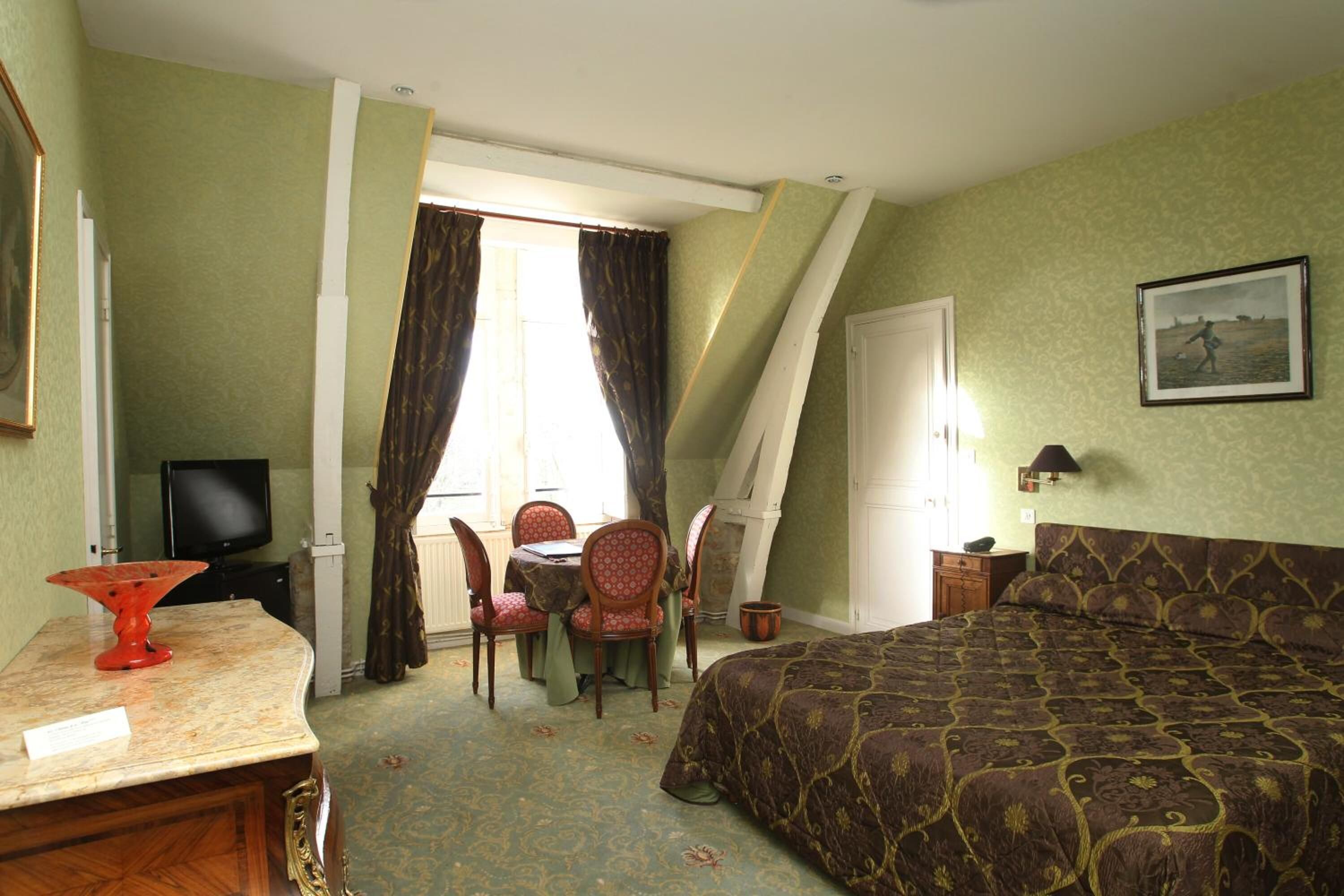 A hotel room with a bed, a desk with a lamp, a television, a dining table with chairs, and a window with curtains.