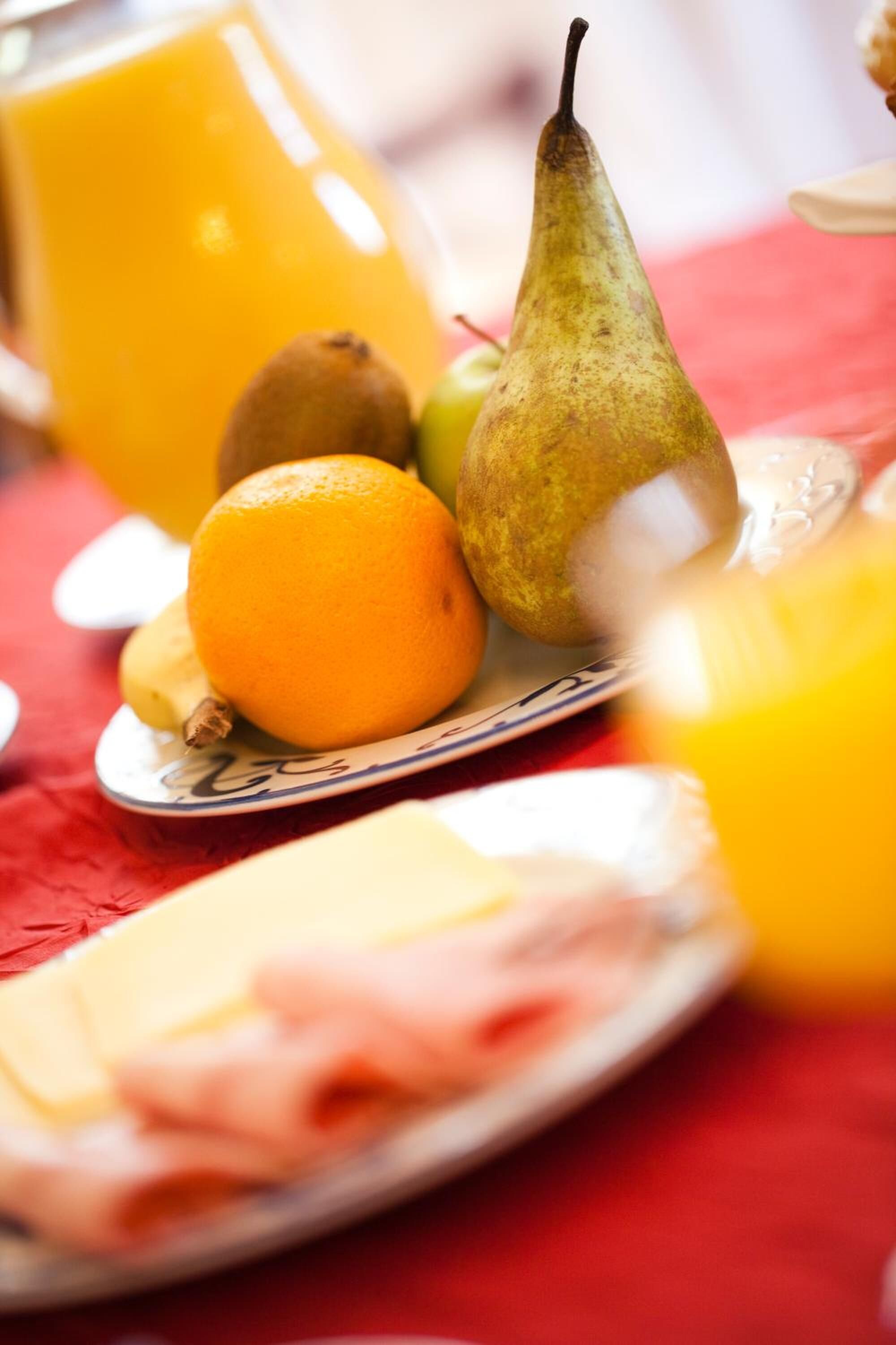 A breakfast setting with a plate of ham, a pear, an orange, and a glass of orange juice on a red tablecloth.
