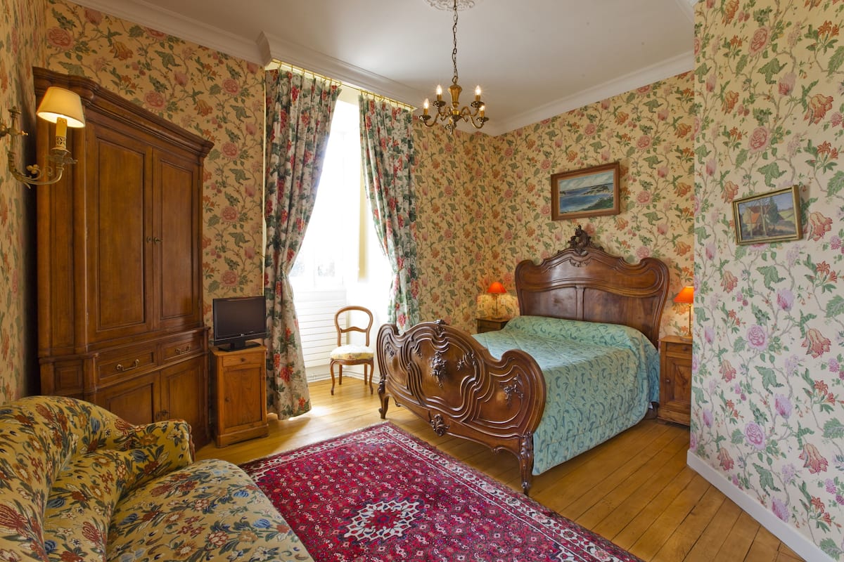 Ein Schlafzimmer mit floralem Wandpapier, einem Holzschrank, einem Bett mit Kopfteil, einem roten Teppich und einem Fernseher auf einer Holzkommode.