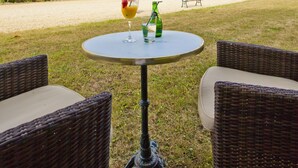 Un patio con mobiliario de mimbre y una mesa con bebidas, un edificio grande con múltiples ventanas y un jardín al fondo.