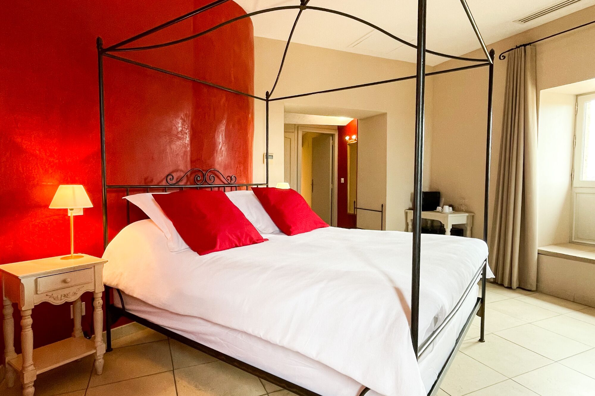 A four-poster bed with white and red bedding, a bedside table with a lamp, and a view of a hallway and window.
