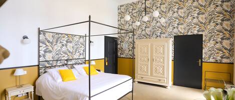 A four-poster bed with white bedding and yellow pillows, a bedside table with a lamp, a wardrobe with a mirror, and a floral wallpaper pattern on the walls.