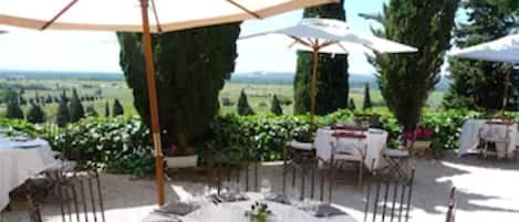 Un área de comedor al aire libre con mesas redondas, manteles blancos y sillas de metal.