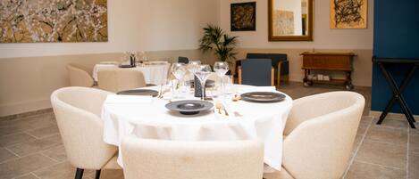 A dining area with a round table set for four, surrounded by beige chairs. There are artworks on the walls and a potted plant in the background.
