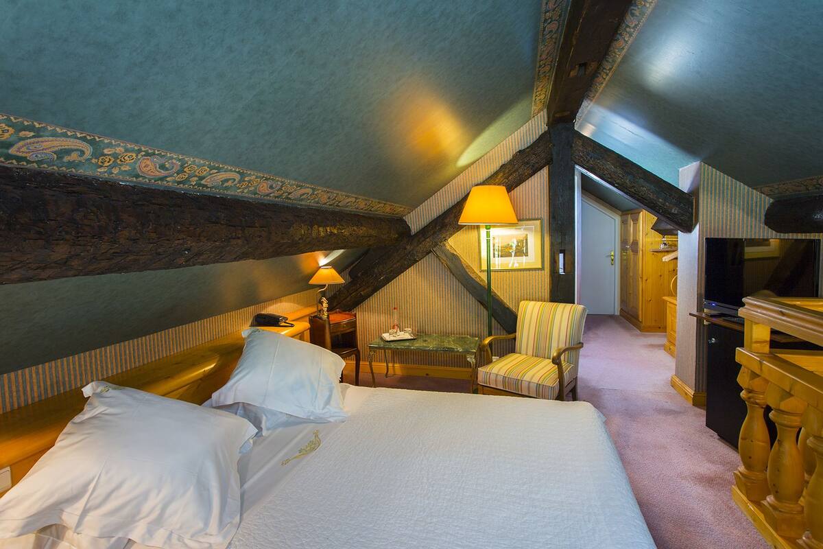 Une chambre avec un grand lit, une chaise, une table et une lampe. Un escalier mène à une autre pièce.