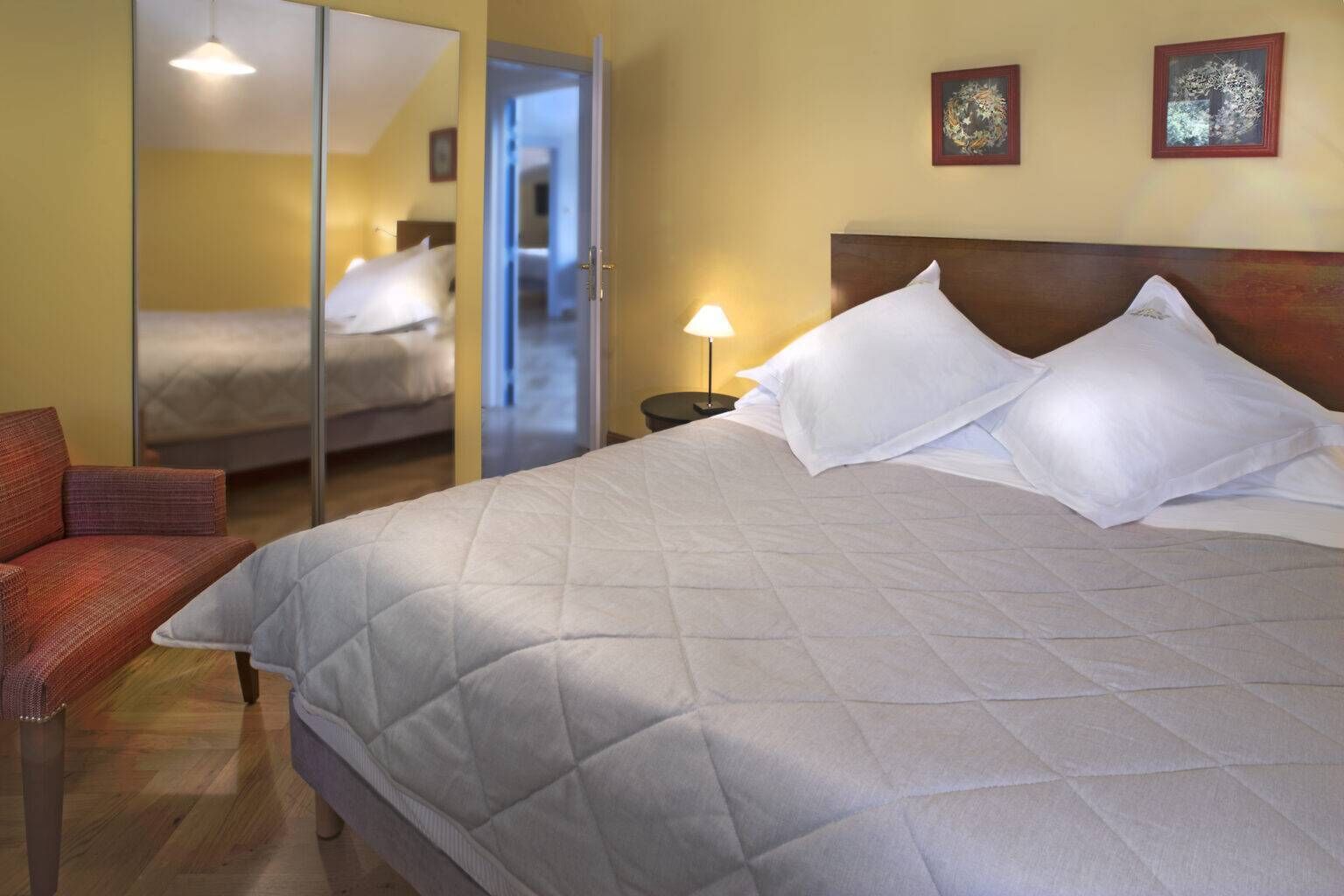 Een hotelkamer met een bed, een stoel, een nachtkastje met een lamp en een deur naar een andere kamer.