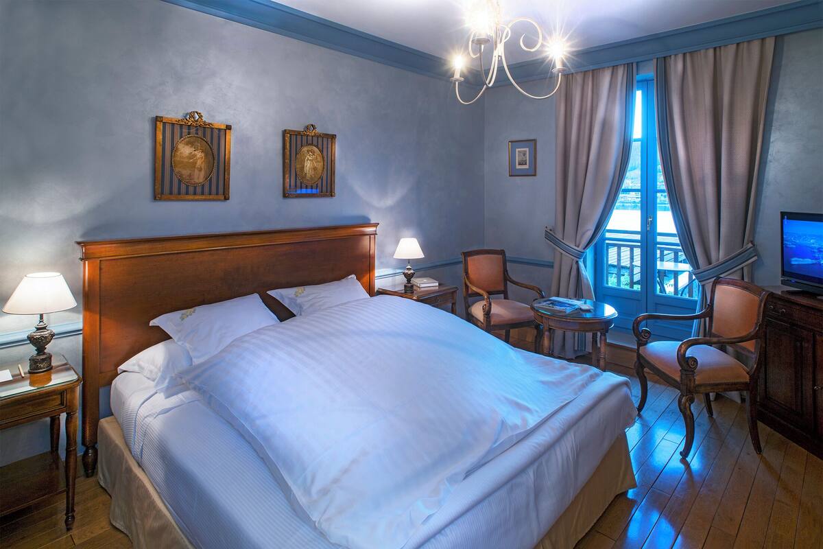 Une chambre d’hôtel avec un grand lit, un bureau avec une lampe, deux chaises, une télévision et une fenêtre avec des rideaux.