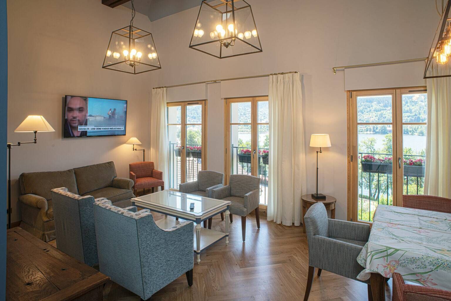 A spacious living room with a sofa, chairs, a coffee table, and a TV. Large windows offer views of greenery outside.