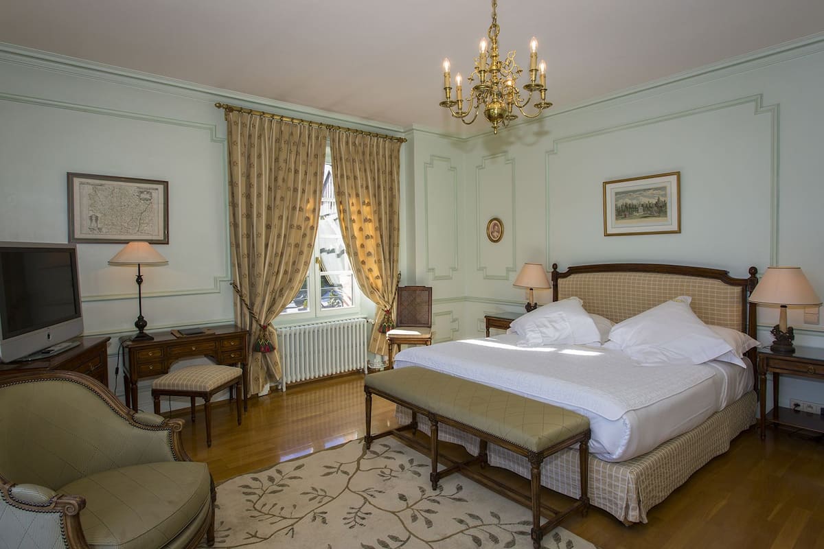 Une chambre d’hôtel classique, dotée d’un grand lit, de deux tables de chevet avec des lampes, d’une télévision, d’un fauteuil et d’une fenêtre avec des rideaux.