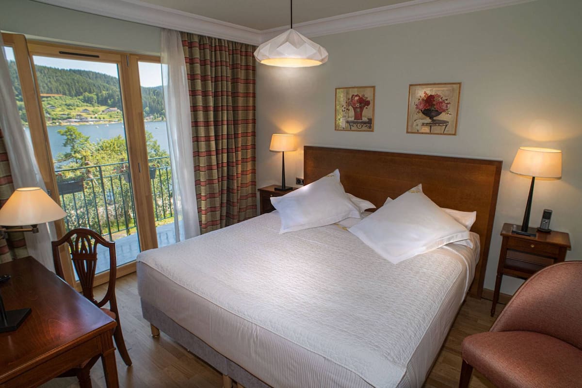 Une chambre d’hôtel avec un grand lit, deux lampes de chevet, une tête de lit en bois, un bureau avec une chaise et une vue sur un lac, visible à travers une fenêtre munie de rideaux.