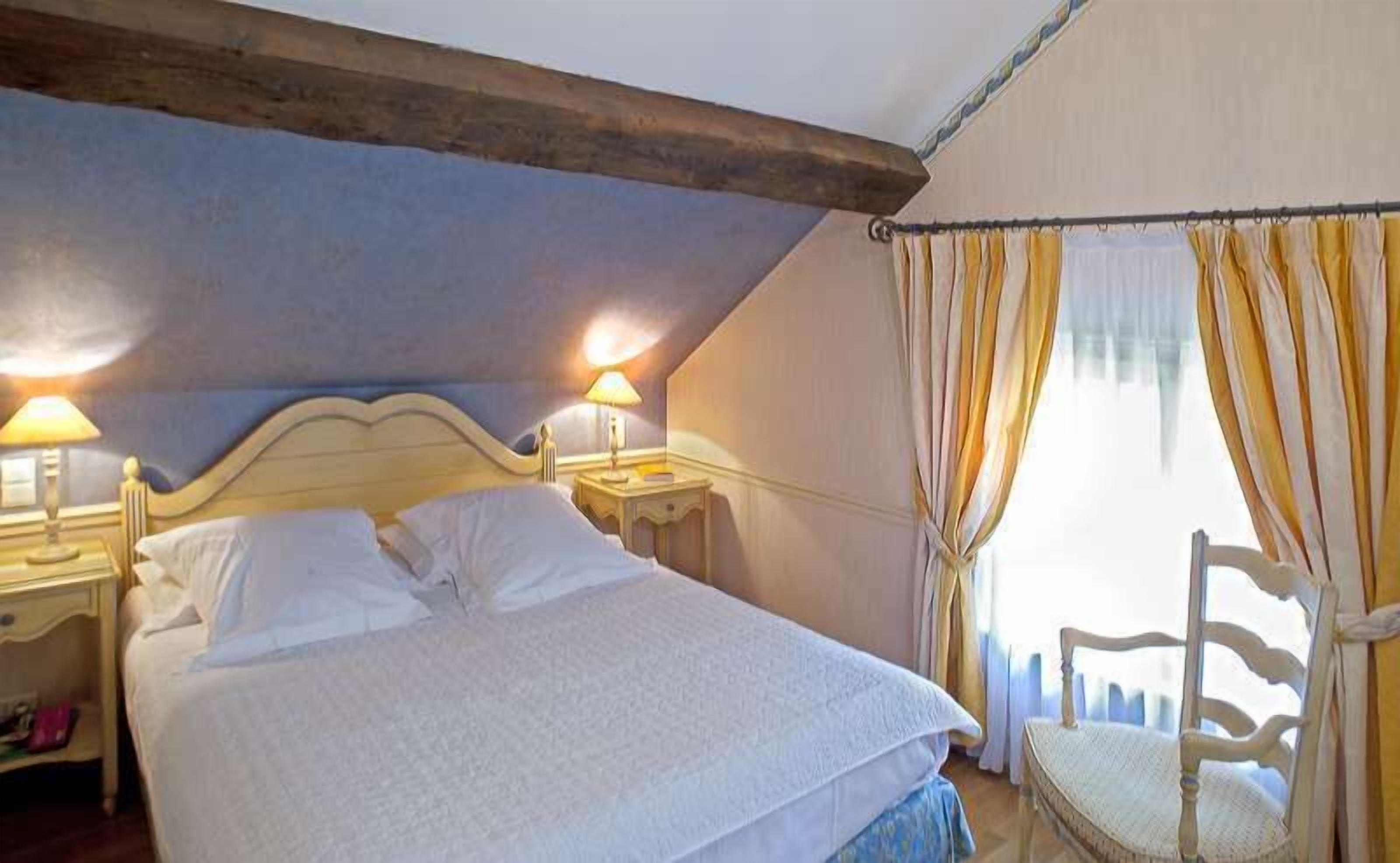 A bedroom with a bed, two bedside lamps, a chair, and a window with curtains.