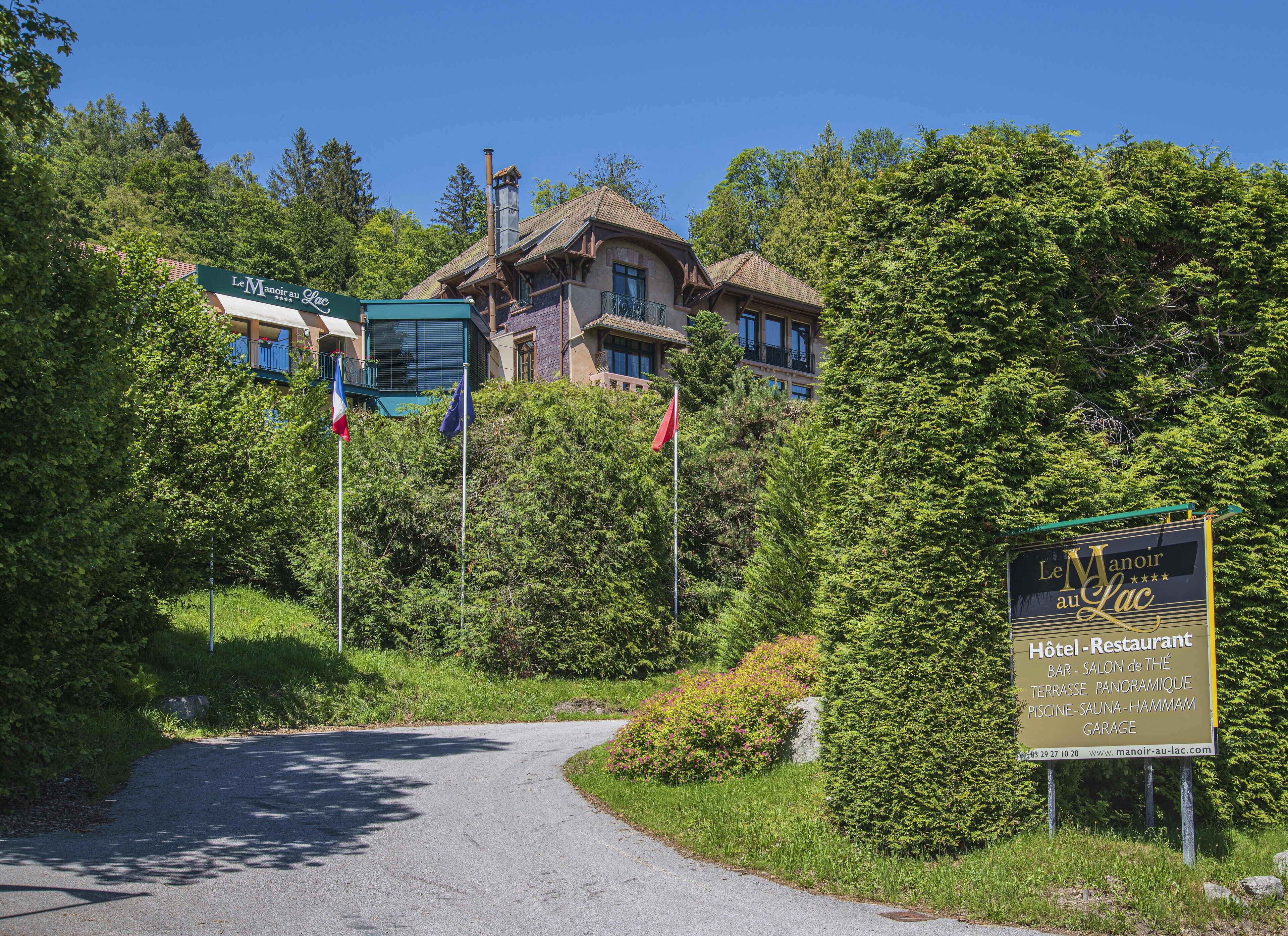 A sign for Le Manoir au Lac Hotel-Restaurant with a scenic view of a building surrounded by lush greenery.