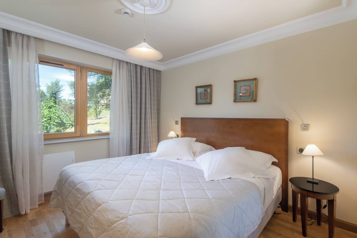 Un lit bien fait, avec du linge de lit blanc, dans une pièce dotée d’une grande fenêtre, d’une tête de lit en bois et de deux lampes fixées au mur.