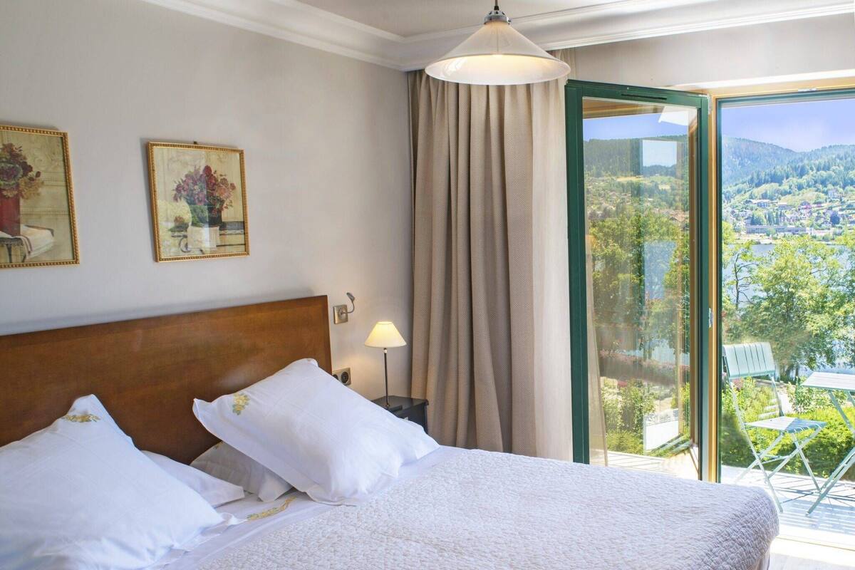 Une chambre d’hôtel avec un grand lit, une table de chevet avec une lampe, une fenêtre donnant sur la ville et les collines, et deux tableaux encadrés accrochés au mur.