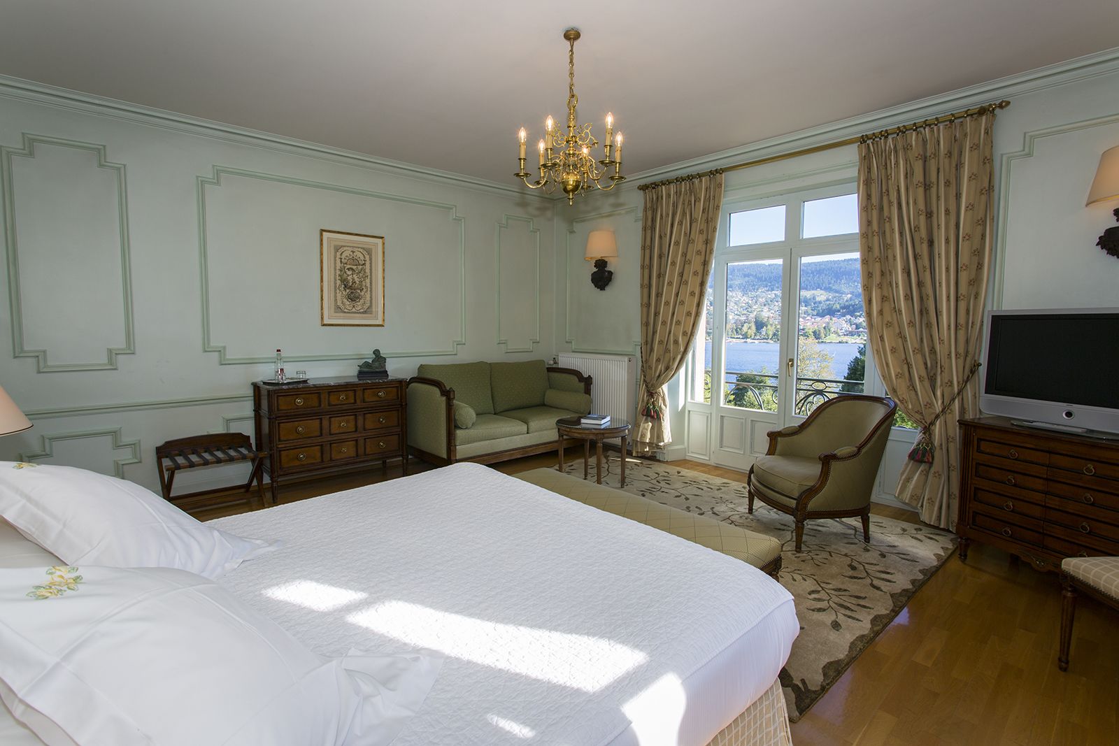 Ein Hotelzimmer mit einem großen Bett, einem Sofa, einem Sessel, einem Schminktisch und Blick auf einen See durch das Fenster.