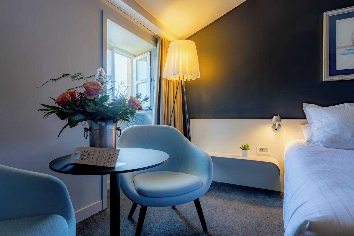 A modern hotel room with a bed, a desk with a vase of flowers, a chair, and a lamp.