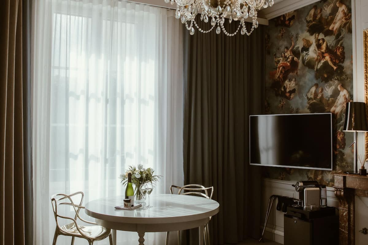 A hotel room with a large bed, a round dining table with chairs, a chandelier, a flat-screen TV, and a painting on the wall.