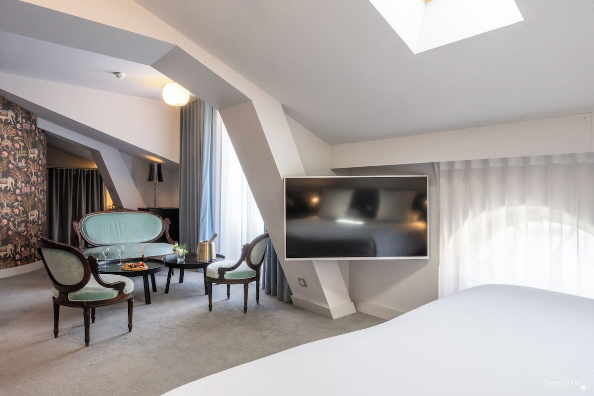 A modern hotel room with a large bed, a flat-screen TV, two armchairs, a small table with a drink, and a decorative wall.