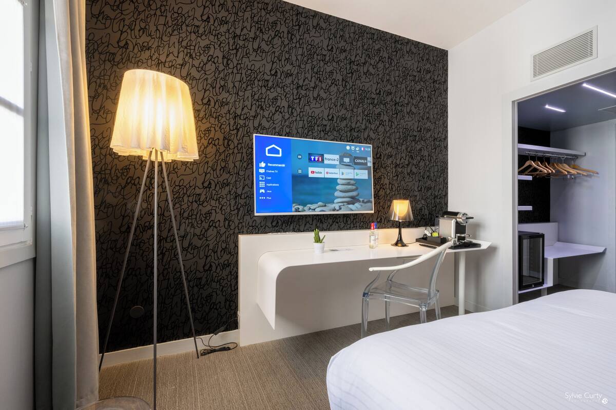 A modern hotel room with a white desk, a large flat-screen TV mounted on the wall, a bedside table with a lamp, a bed, and a wardrobe with hanging clothes.