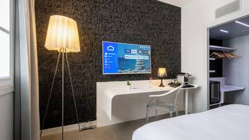 Una habitación de hotel moderna con un escritorio blanco, un televisor de pantalla plana grande montado en la pared, una mesita de noche con lámpara, una cama y un armario con ropa colgada.