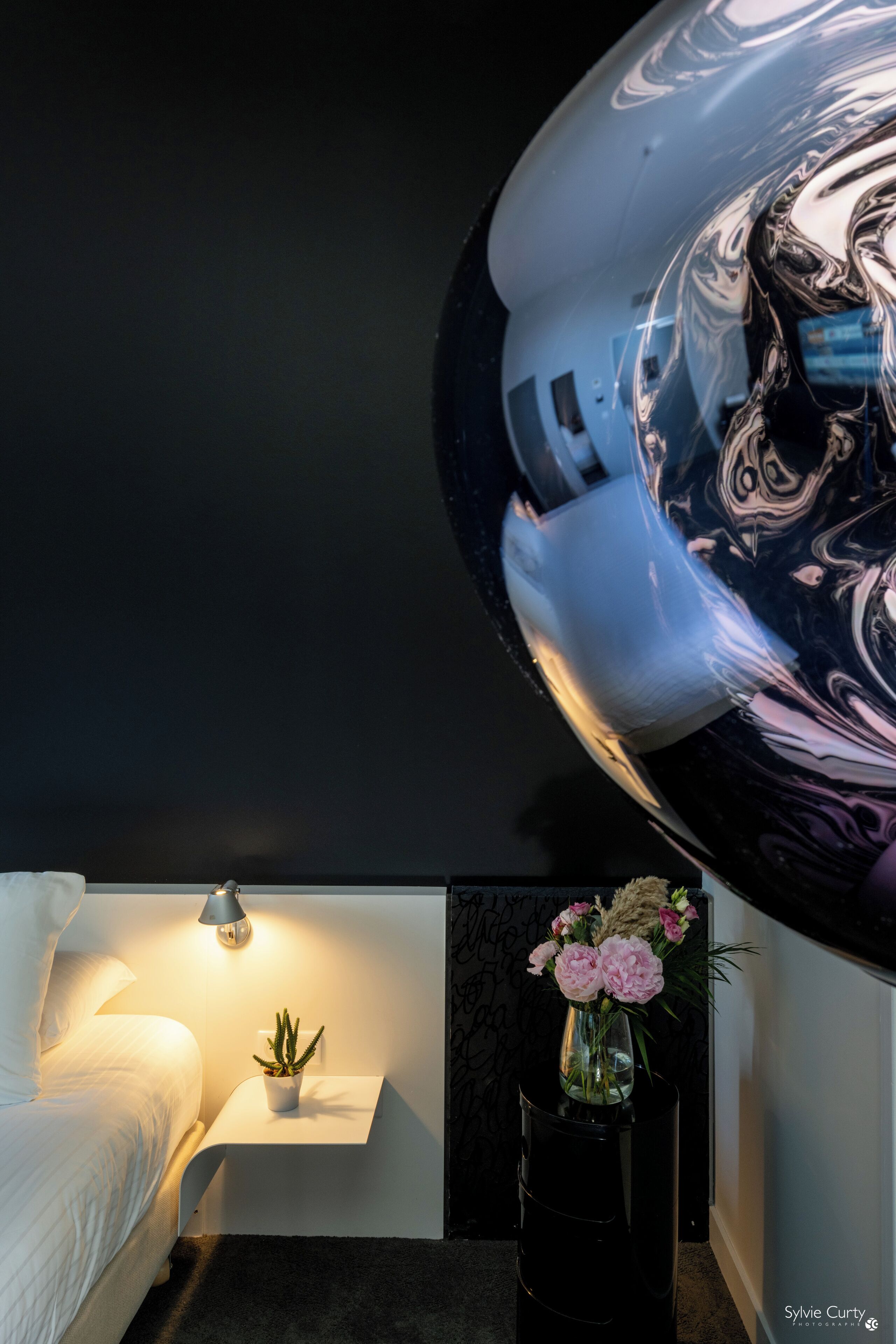 A modern bedroom with a white bed, a small bedside table with a lamp, a vase with flowers, and a large mirror reflecting the room.