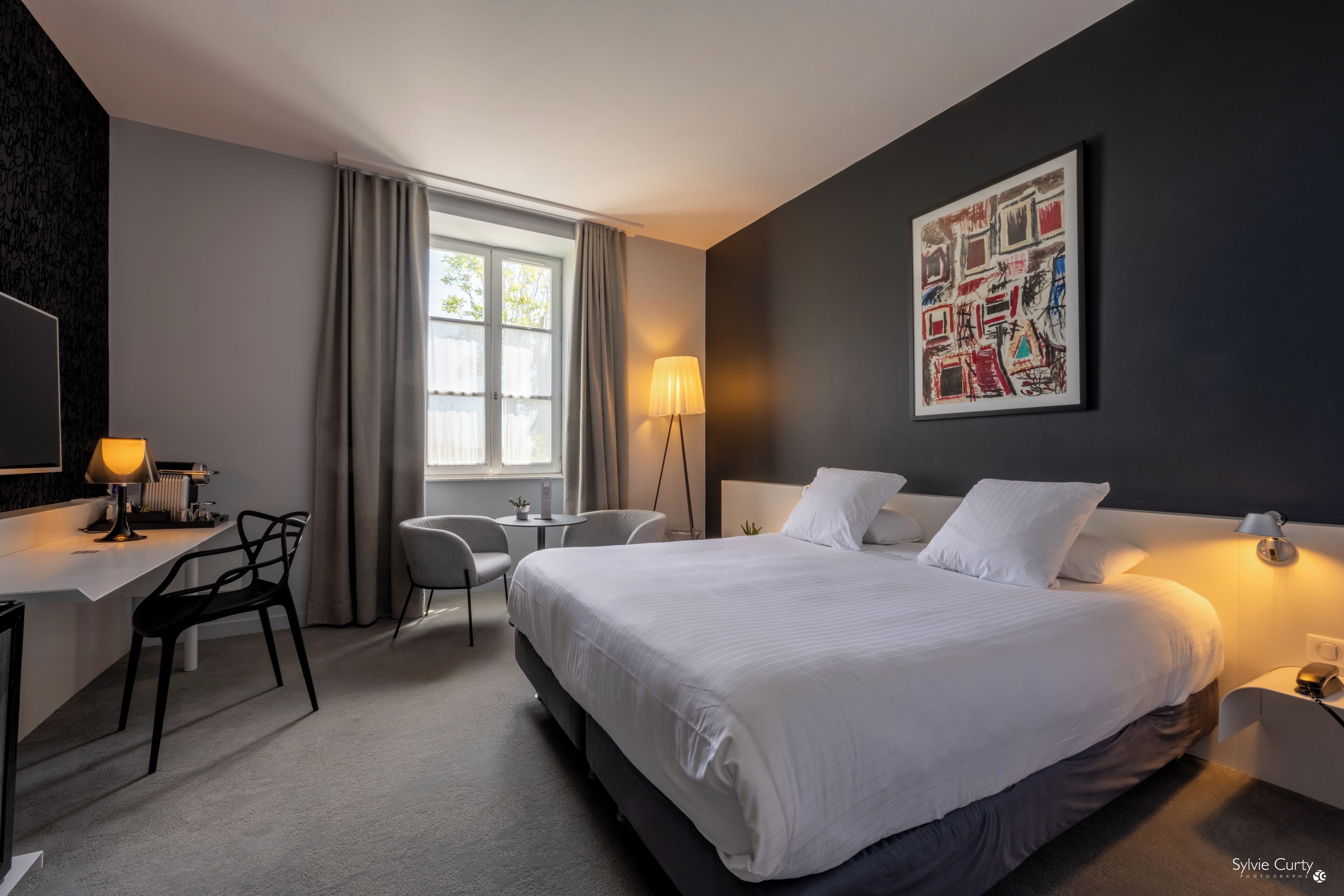 A modern hotel room with a large bed, a desk, a chair, and a window with curtains.