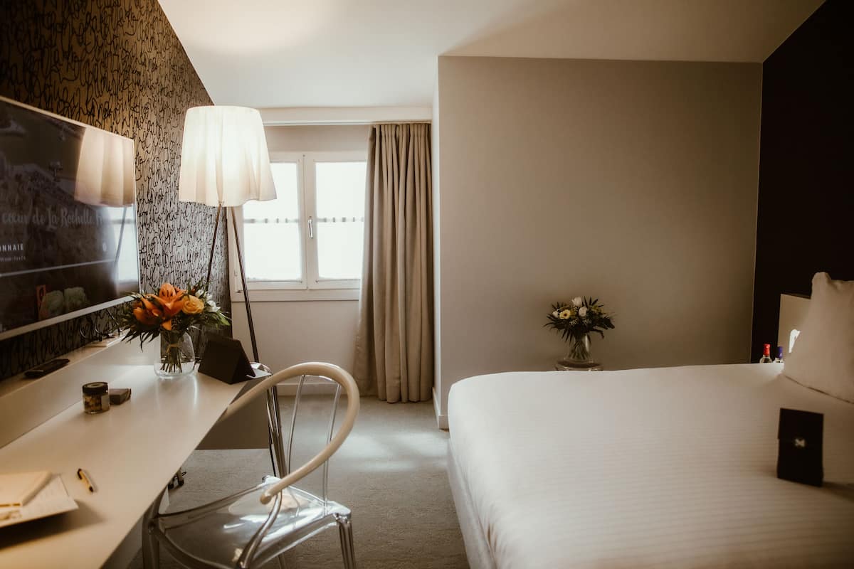 A hotel room with a bed, a desk with a lamp, a chair, a TV, and a window with curtains.
