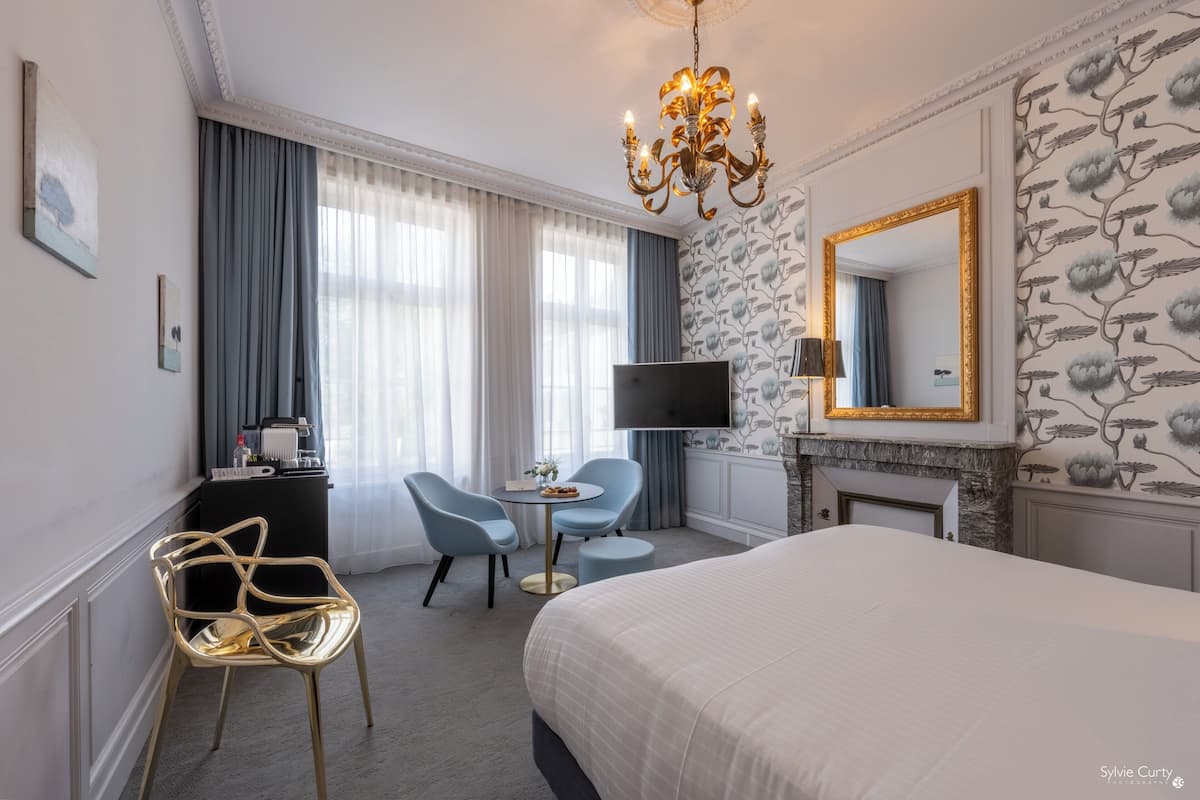 A hotel room with a large bed, a TV, a desk with a chair, a fireplace, and a chandelier.