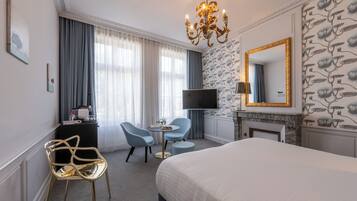 A hotel room with a large bed, a TV, a desk with a chair, a fireplace, and a chandelier.