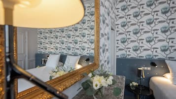 Habitación de hotel con un espejo grande, una cama con ropa blanca, un florero con flores, una mesita con una lámpara y papel tapiz floral.