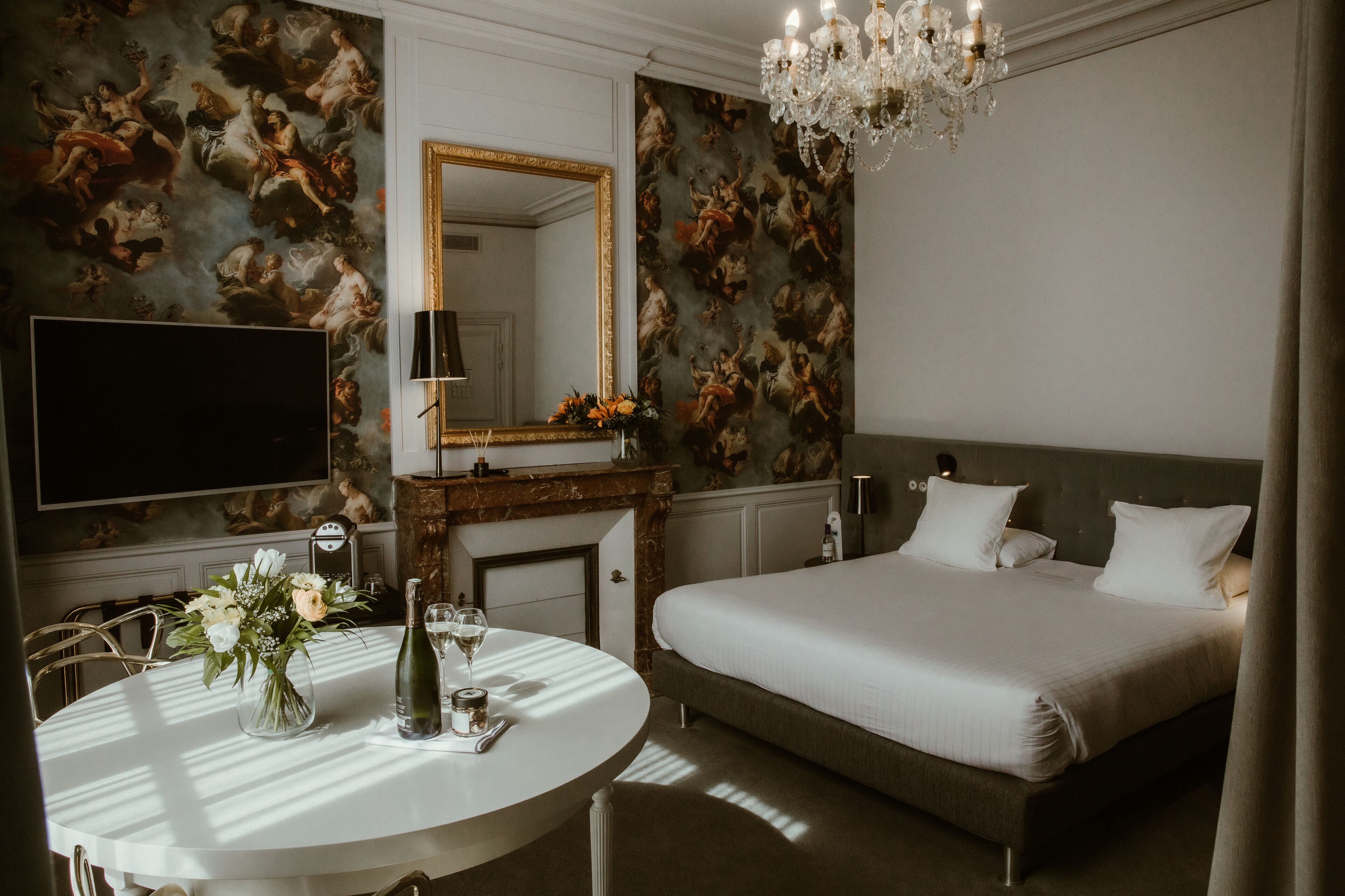 Una habitación de hotel con una cama grande, una mesa redonda con un jarrón de flores, una chimenea y un espejo amplio.