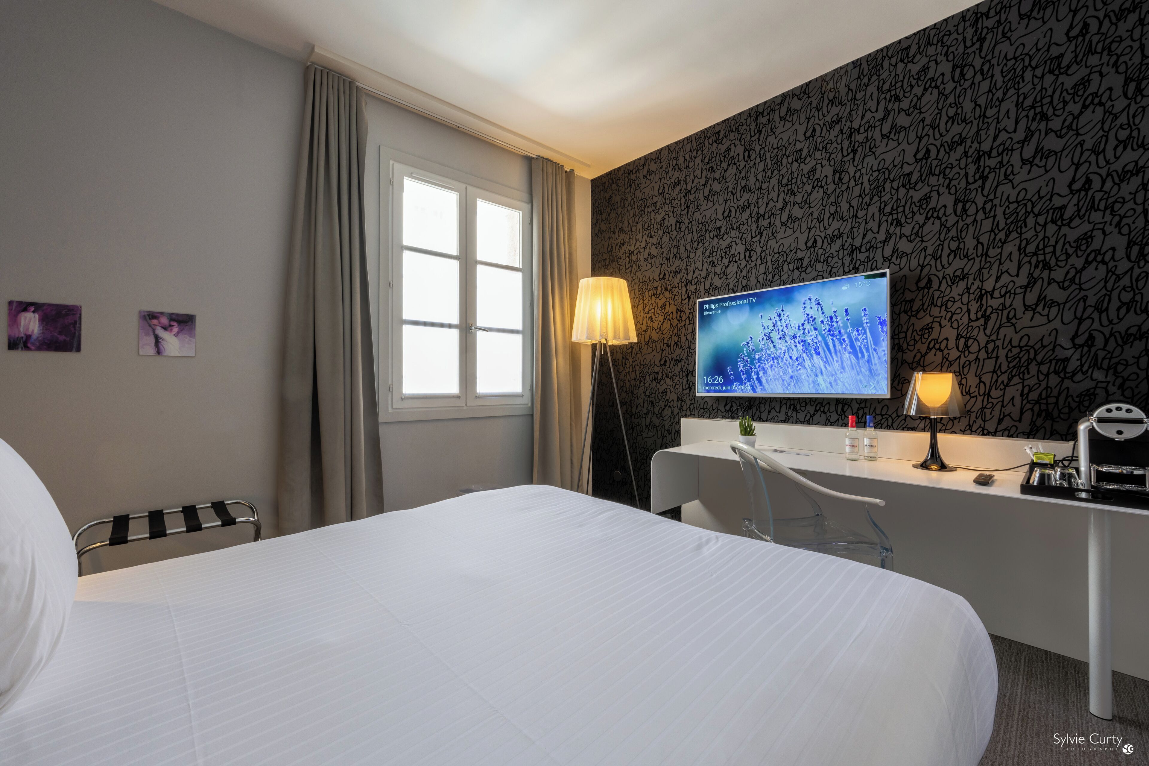 A hotel room with a bed, a desk with a computer, a lamp, and a TV displaying a nature scene.
